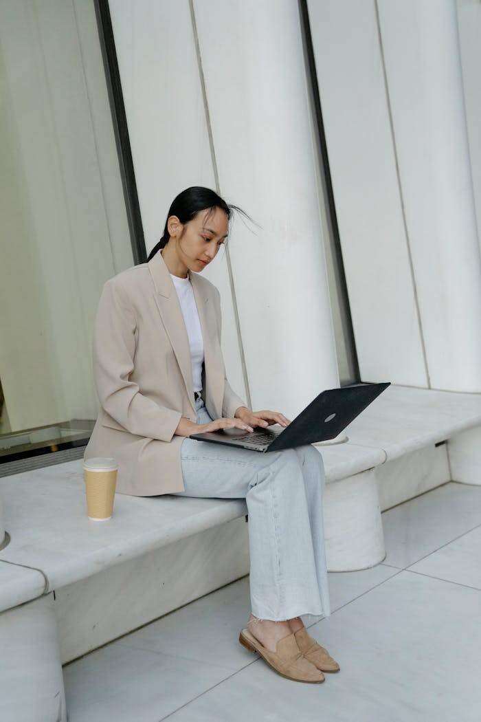 Asian businesswoman sitting outdoors using a laptop with a coffee to go.